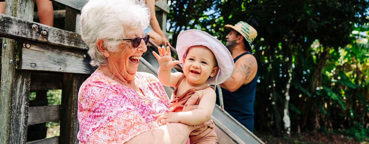 Elderly woman holding her young grandchild. They're both on a playground smiling in the sunshine.