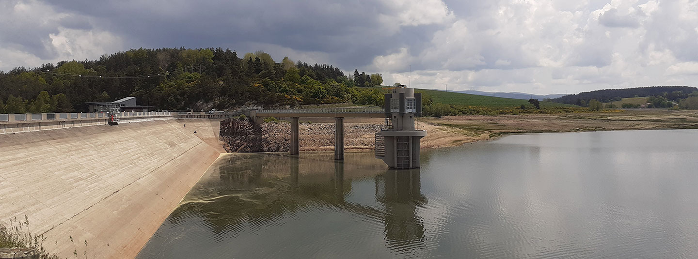 Barrage en béton sur un plan d’eau calme, avec une tour de contrôle reliée par une passerelle, des collines boisées à l’arrière-plan et un ciel partiellement nuageux.