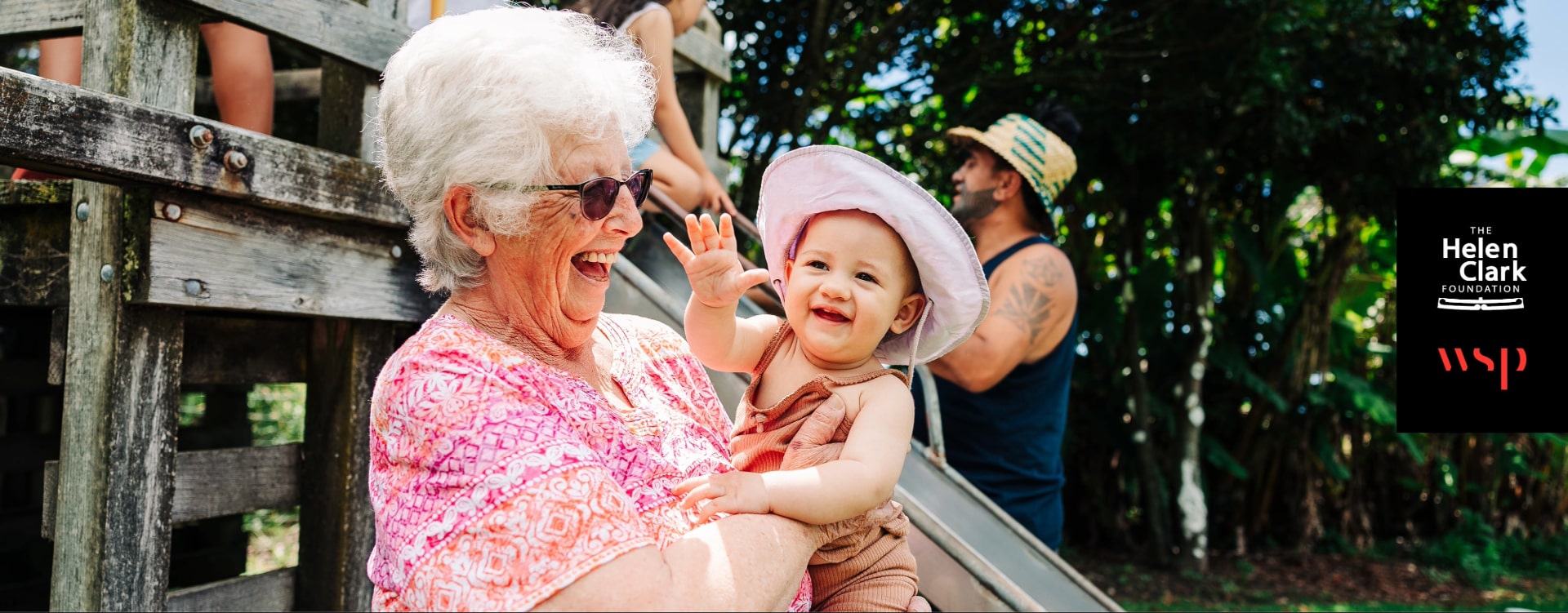 Elderly woman holding her young grandchild. They're both on a playground smiling in the sunshine.