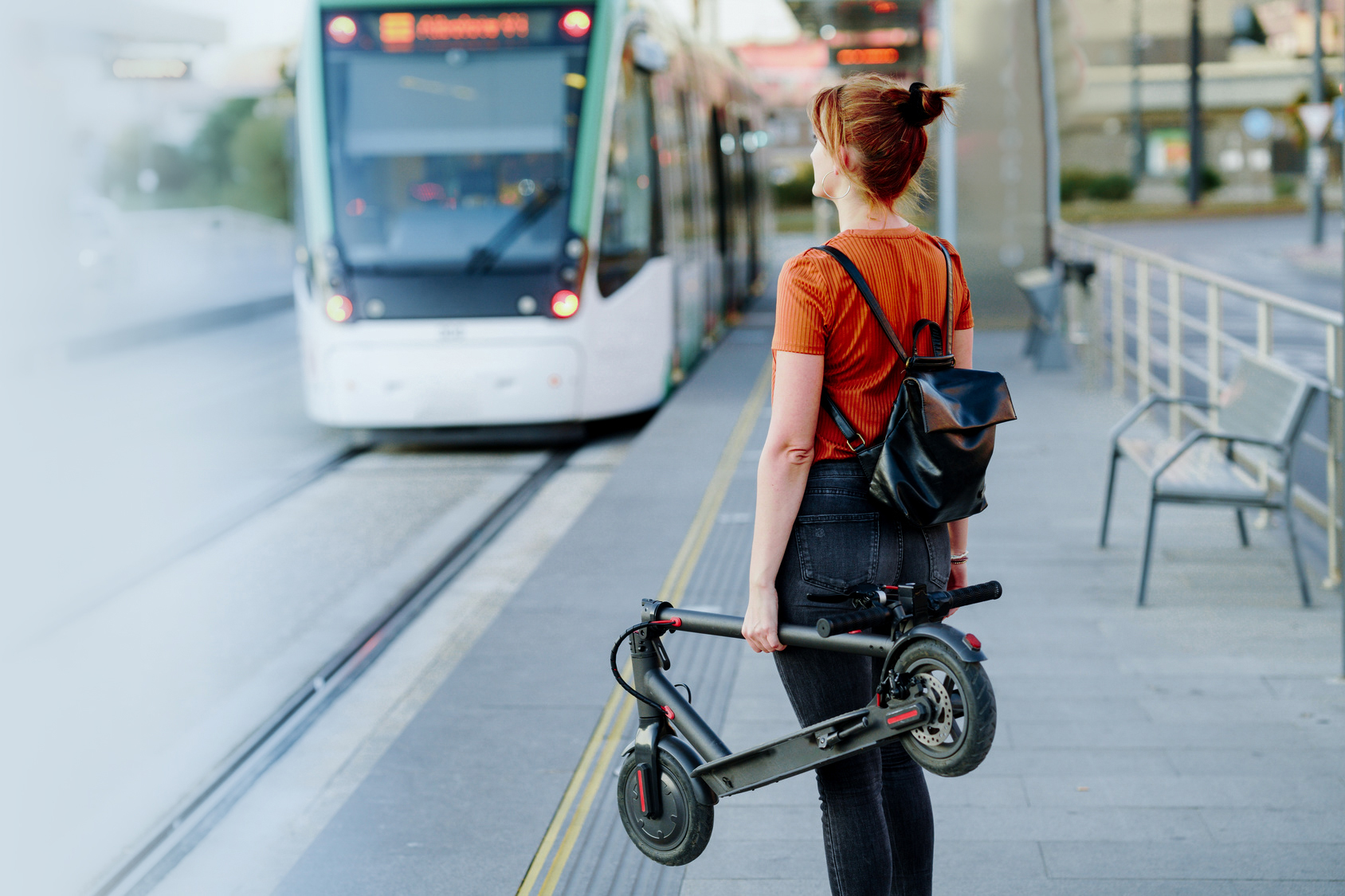 Back-view-of-woman-with-backpack-and-folded-electric-scooter-waiting-at-tram-stop