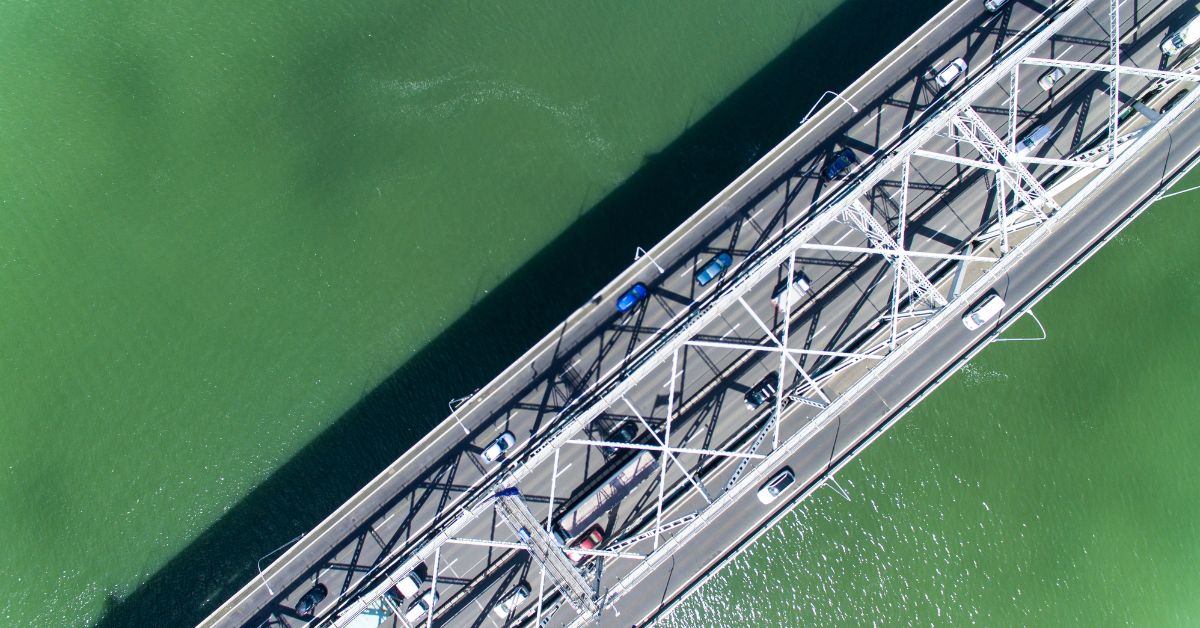 Drone shot looking straight down onto the Auckland Harbour Bridge road. There are cars commuting both ways across the bridge. The top left corner is dominated by a stretch of the Waitemata Bay blue-green water. 