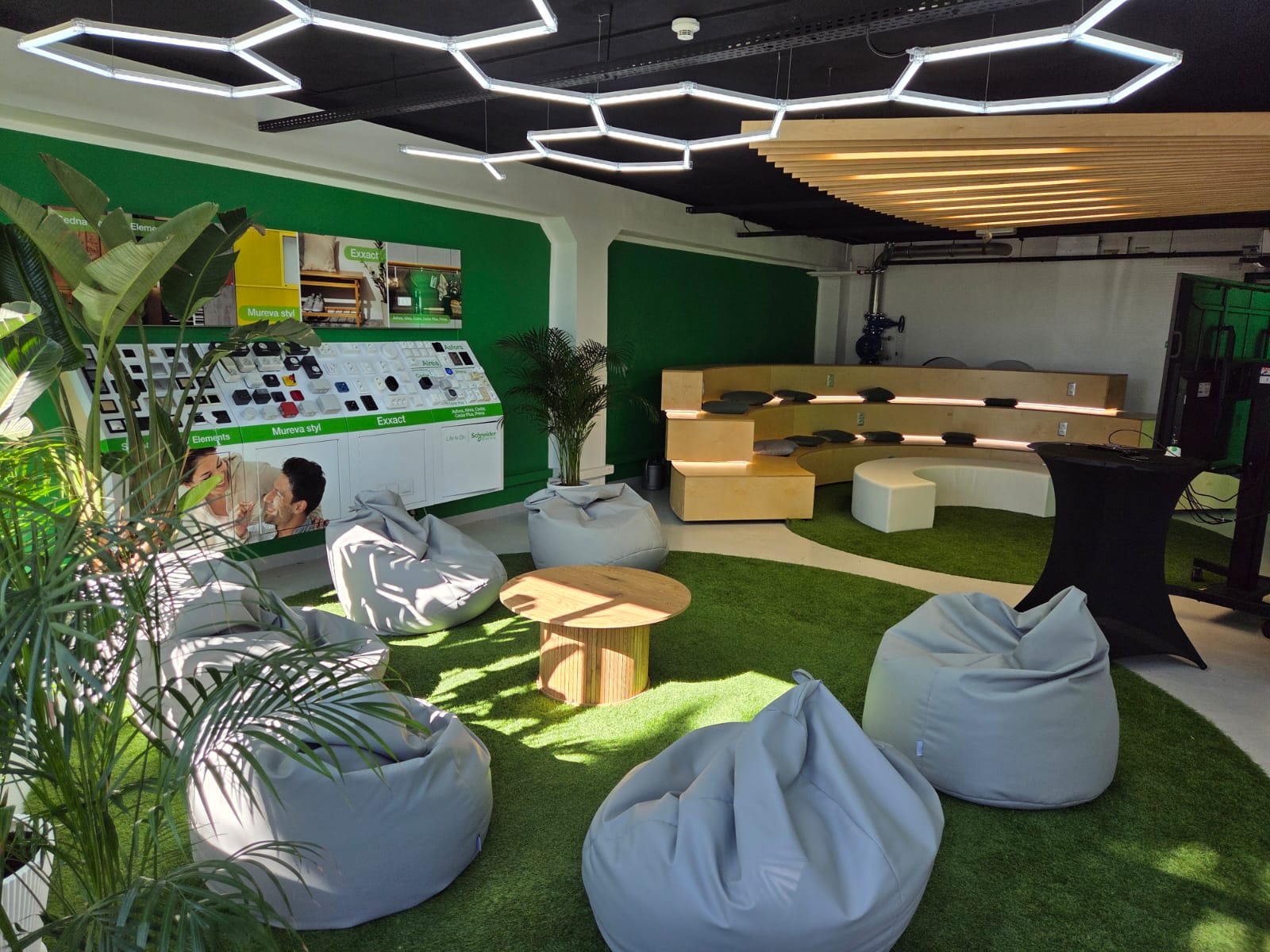 Espace de collaboration moderne avec sol en gazon synthétique, poufs, gradins en bois éclairés, plantes d’intérieur et luminaires géométriques au plafond.