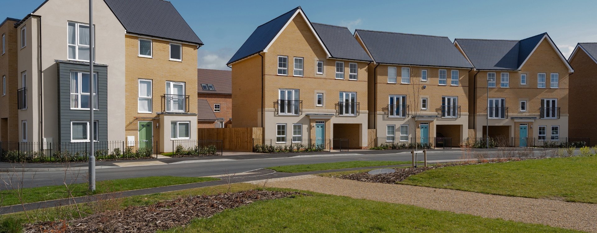 Image showcasing a modern housing estate in the United Kingdom. This includes a series of newly built terraced townhouses. A road runs in front of them and there is also a grassy area in the foreground with newly planted trees. 